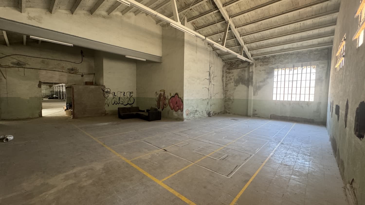 A large empty factory room, concrete floor with painted markings, high timber roof and a barred window
